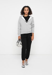 Graue Zip-Hoodie mit lässigem Schnitt, kombiniert mit schwarzen Jogginghosen. Das Modell hält eine kleine weiße Handtasche mit schwarzen Akzenten. Silberne Schuhe.
