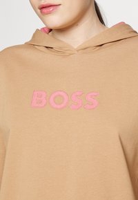 Persona che indossa una felpa beige con un logo ricamato rosa "BOSS" sul petto, mostrando la parte inferiore del viso e il collo.