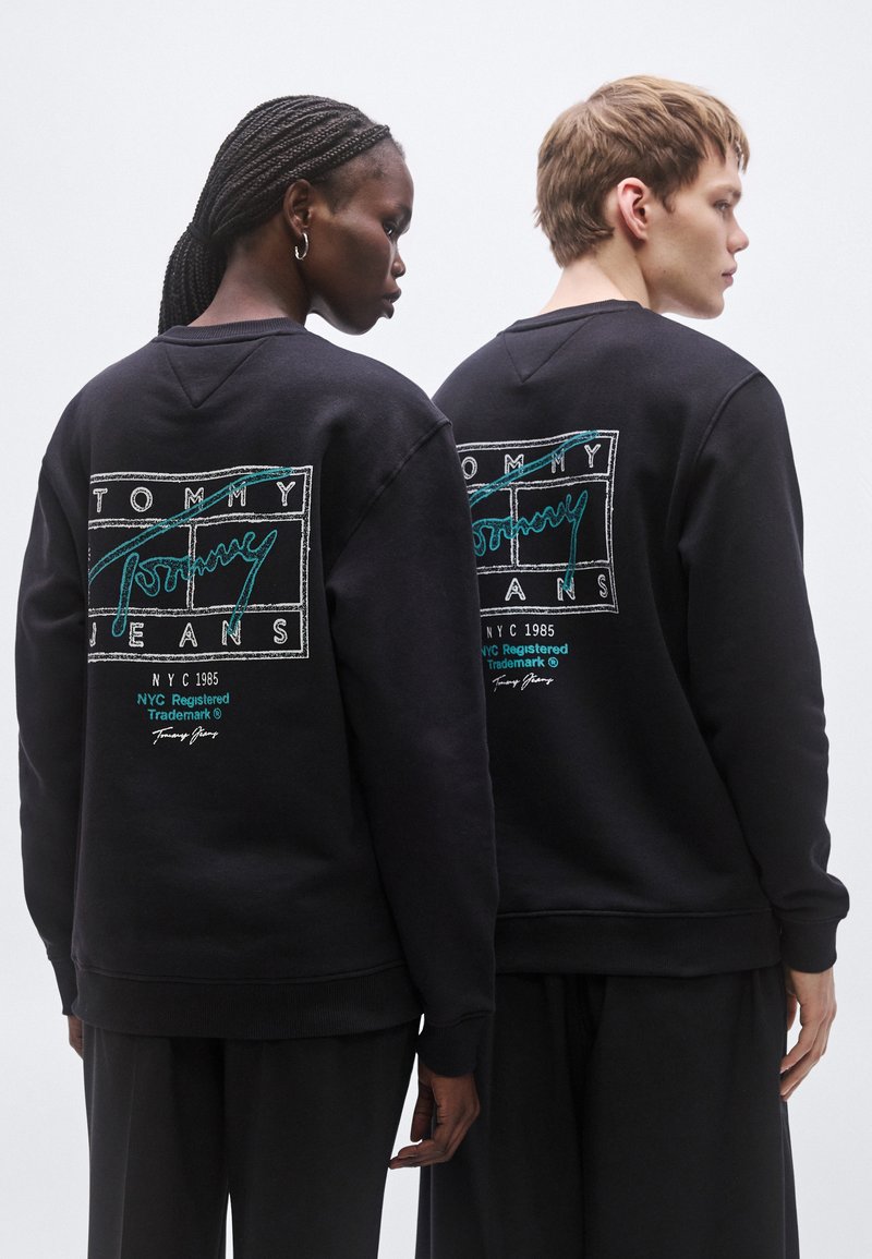 Black sweatshirt featuring a colorful "TOMMY JEANS" logo on the back, with textured fabric and a relaxed fit.