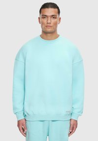 Světle modrý crewneck svetr vyrobený z bavlněné směsi. Má spadlé ramena, žebrované manžety a decentní logo štítek poblíž lemu.