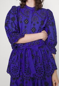 Haut en tissu violet avec broderie florale noire, manches bouffantes, devant à boutonnière et taille nouée pour une silhouette ajustée.