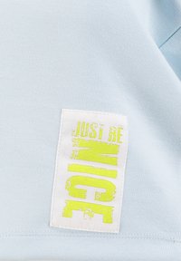 Tela azul claro con un parche blanco cosido que presenta texto amarillo desgastado "JUST BE NICE" en disposición vertical.