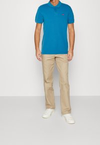 Hombre vistiendo una camiseta polo de manga corta azul, pantalones beiges y zapatillas blancas, de pie contra un fondo blanco liso.