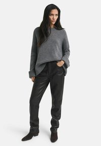 Grauer Strickpullover mit lockerer Passform und breiten Bündchen, kombiniert mit schwarzen Lederhosen und dunklen Knöchelstiefeln. Heller, einfarbiger Hintergrund.