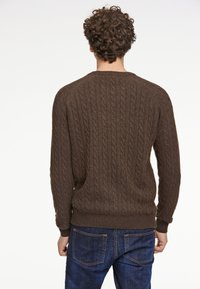 Ruskea palmikkoneule, jossa on ribbireunus kauluksessa ja hihoissa. Teksturoitu kuvio kulkee pystysuoraan, yhdistettynä tummansinisiin farkkuihin.