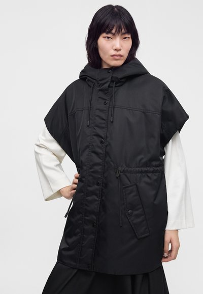 Femme portant un gilet cape à capuche noir par-dessus un haut blanc à manches longues, debout avec une main sur la hanche devant un fond uni.