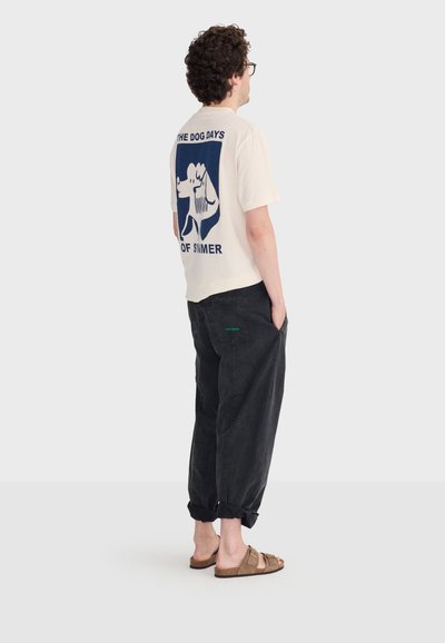Homme aux cheveux bouclés et lunettes portant un T-shirt crème avec un motif de chien bleu et le texte "Les jours caniculaires de l'été", un pantalon noir et des sandales marron.