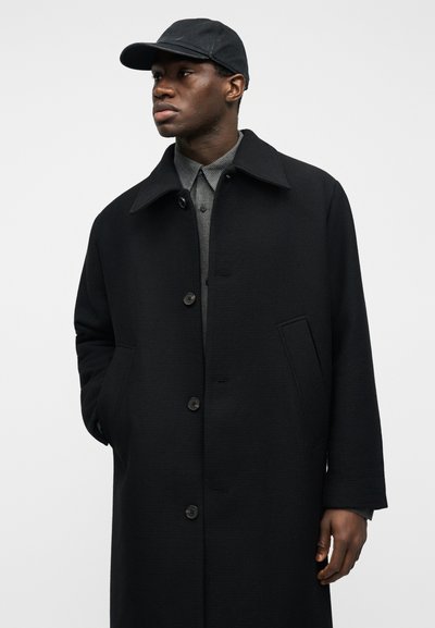 Άνδρας που φοράει μαύρο καπέλο, μαύρο παλτό με κουμπιά και φαρδύ γιακά, και γκρι πουκάμισο με σχέδια, κοιτάζοντας προς τα αριστερά σε απλό φόντο.