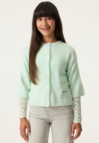 Cárdigan de lana verde claro con mangas tres cuartos, cierre de botones en el frente y detalle bordado en el dobladillo, combinado con jeans grises con lunares.