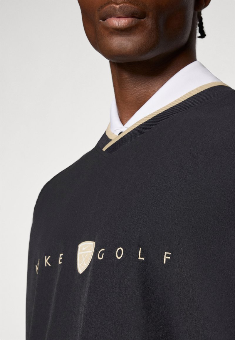 Pull de golf Nike noir avec un col en V, des bordures dorées et un accent logo sur le devant. Texture lisse ; col visible sous le pull.