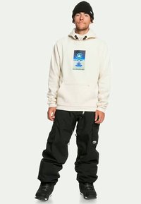 Krém színű kapucnis pulóver kék örvényszerű grafikával, fekete bő nadrággal és fekete hócipővel viselve. A megjelenést egy fekete sapka egészíti ki.