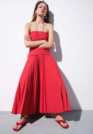 Joven mujer está de pie junto a una pared blanca, lleva una falda plisada roja sin tirantes y sandalias a juego, con los brazos cruzados, cabello largo y pendientes de aro.