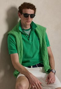 Mladý muž s tmavými slnečnými okuliarmi, zelenou polokošeľou, zeleným svetrom prehodeným cez plecia, bielymi krátkymi nohavicami a hodinkami na zápästí, sediaci oproti béžovej stene.
