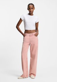 Valkoinen lyhennetty t-paita ja vaaleanpunaiset leveälahkeiset farkut, joissa on repsottavat reunat. Malli poseeraa kädet taskuissa yksiväristä taustaa vasten.