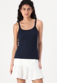 Débardeur bleu marine, design ajusté, bretelles fines, texture côtelée. Porté avec une jupe blanche à volants, style minimaliste.