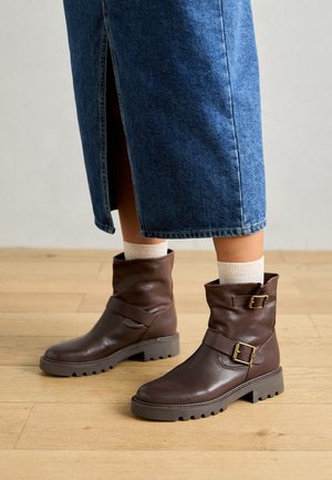 Barna bőr bokacsizmák, laza dizájnnal, csattal díszítve és texturált gumitalppal. Világos színű zoknikkal és farmer szoknyával viselve.