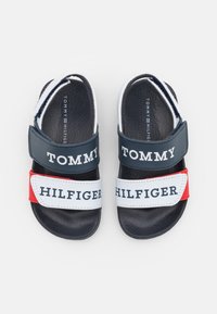 Navy, punainen ja valkoinen sandaalit tarranauhalla. Liuskoissa on "TOMMY" ja "HILFIGER" -logot. Teksturoitu musta pohja.