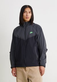 Nike vetrovka v temno sivi in črni barvi, z visokim ovratnikom, zadrgo in majhnim zelenim logotipom. Lahka in s teksturiranim zaključkom.
