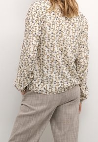 Kwiatowa bluzka w kremowym kolorze z niebieskimi i brązowymi wzorami; długie rękawy, luźny krój. Połączona z beżowymi spodniami w kratę z delikatną teksturą.