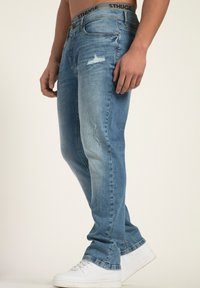 Jeans de denim azul claro con un ajuste relajado, que presentan detalles desgastados en el muslo y una cinturilla negra. Combinados con zapatillas deportivas blancas.
