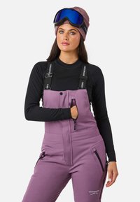 Vrouw in paarse skibroek, zwart langemouwen top, mauve beanie en blauwe skibril, met hand in de rits van de borstzak.