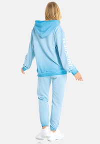 Hellblaue Kapuzenjacke und passende Jogginghose aus weichem Baumwollstoff, mit einer großen Kapuze und dem Text "UNIVERSAL" auf dem Ärmel. Weiße Sneakers.