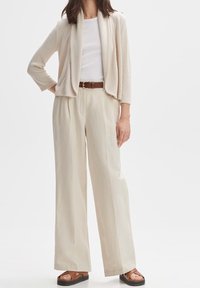 Jasno beżowy kardigan z luźnym przodem, długimi rękawami, zestawiony z kremowymi spodniami o szerokich nogawkach i brązowymi skórzanymi sandałami.