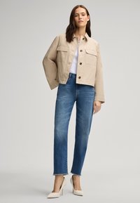 Giacca cropped beige con due tasche sul petto, abbinata a jeans azzurri a gamba dritta e scarpe con tacco beige.