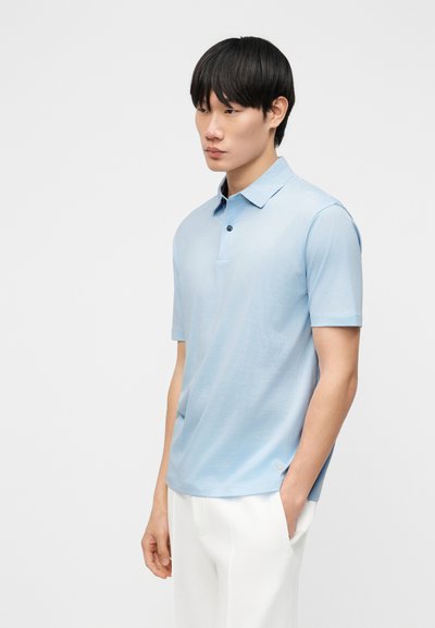 Νέος άνδρας με μαύρα μαλλιά, ντυμένος με ανοιχτό μπλε πόλο κοντομάνικο και λευκό παντελόνι, στέκεται με το ένα χέρι στην τσέπη.