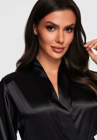 Donna con lunghi capelli scuri che indossa un accappatoio di satin nero, sorridendo delicatamente, toccandosi i capelli vicino all'orecchio destro su uno sfondo semplice.