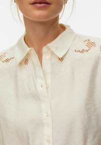 Blouse couleur crème avec col pointu, fermeture boutonnée et détails en dentelle sur les épaules. Texture douce et froissée sur toute la surface.