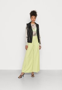 Jaqueta preta de pele sem mangas, blusa transparente com padrão, e calças largas amarelo-lima combinadas com sandálias pretas com tiras.