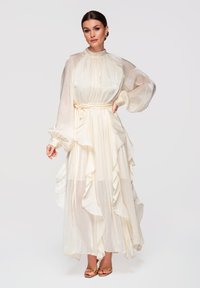 Femme portant une longue robe crème fluide avec des manches longues bouffantes et des détails à volants, assortie à des talons ouverts dorés, debout sur un fond blanc.