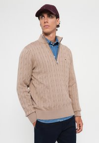 Jonge man in een beige kabelgebreide trui met rits over een blauwe denimshirt en donkere broek, met een kastanjebruine pet, die opzij kijkt.