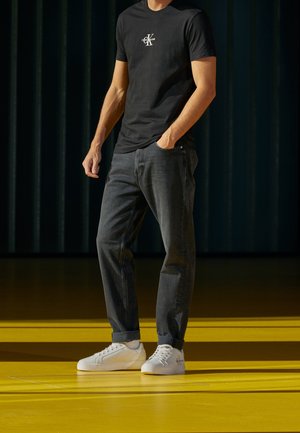 Mężczyzna stoi na żółtej podłodze, ubrany w czarną koszulkę Calvin Klein, ciemne jeansy z mankietami, białe trampki, jedną ręką w kieszeni, ciemne tło.