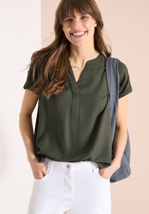 Jeune femme souriante, portant un chemisier vert foncé à col en V et un pantalon blanc, avec un sac gris porté sur une épaule.