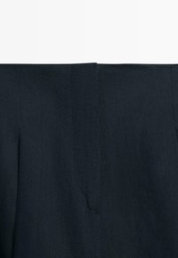 Gros plan sur un tissu bleu marine foncé avec des coutures verticales visibles et un détail subtil de couture, probablement une partie de la ceinture d'un vêtement.