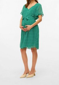 Vestido de maternidad verde con puntos, de manga corta, escote en V y cintura ajustable. Modelo luciendo mules beige de punta.