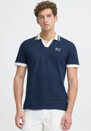 Junger Mann mit lockigem Haar, der ein marineblaues kurzärmeliges Polohemd mit weißem Rand trägt und vor einem schlichten hellen Hintergrund steht.