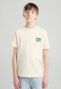 T-shirt en coton couleur crème avec des manches courtes, mettant en avant le texte vert "GOOD LIFE" sur le côté gauche de la poitrine. Coupe décontractée, design col rond.