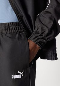 Black athletic jacket with elastic cuffs and a zippered pocket. Features white Puma logo and light blue interior lining. Smooth surface texture.