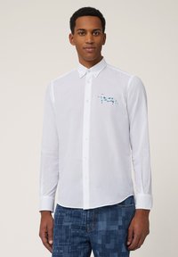 Chemise blanche à manches longues avec un col boutonné et un motif de vache bleue sur la poitrine gauche. Le tissu est lisse avec une coupe classique.