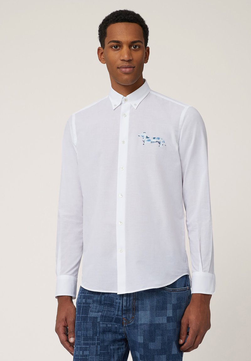 Chemise blanche à manches longues avec un col boutonné et un motif de vache bleue sur la poitrine gauche. Le tissu est lisse avec une coupe classique.