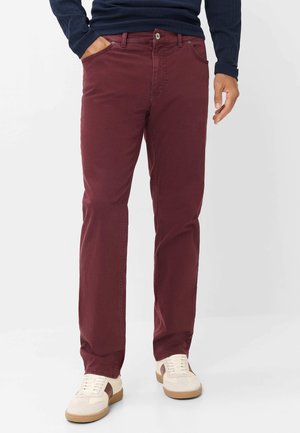 Uomo che indossa pantaloni bordeaux a taglio dritto, camicia blu navy a maniche lunghe e sneakers beige con dettagli marroni, in piedi con una mano in tasca.