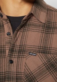 Chemise à carreaux marron et vert foncé boutonnée avec poche avant et étiquette logo Volcom, portée par-dessus un sous-vêtement noir.