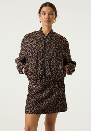 Vrouw staat, draagt een luipaardprint jas met rits en bijpassende rok, handen in de zakken, kijkt vooruit met een neutrale uitdrukking.