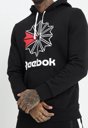 Mężczyzna w czarnej bluzie Reebok z białymi ściągaczami, geometryczne logo w kolorze czerwonym i białym, splecione ręce ukazujące szczegółowe tatuaże.