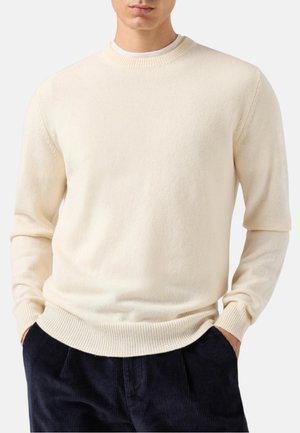 Uomo che indossa un maglione crema semplice con scollo rotondo, polsini e orlo a coste, abbinato a pantaloni di velluto a coste scuro, mani in tasca.