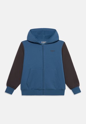 Hoodie azul com fecho e mangas compridas pretas, bolsos frontais e a palavra "TINY" impressa no peito e no interior da gola.