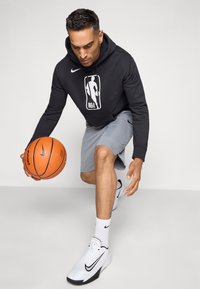 Čierna mikina Nike s logom NBA, šedé športové šortky, biele topánky Nike, drží oranžovú basketbalovú loptu, v pokrčenej pozícii.